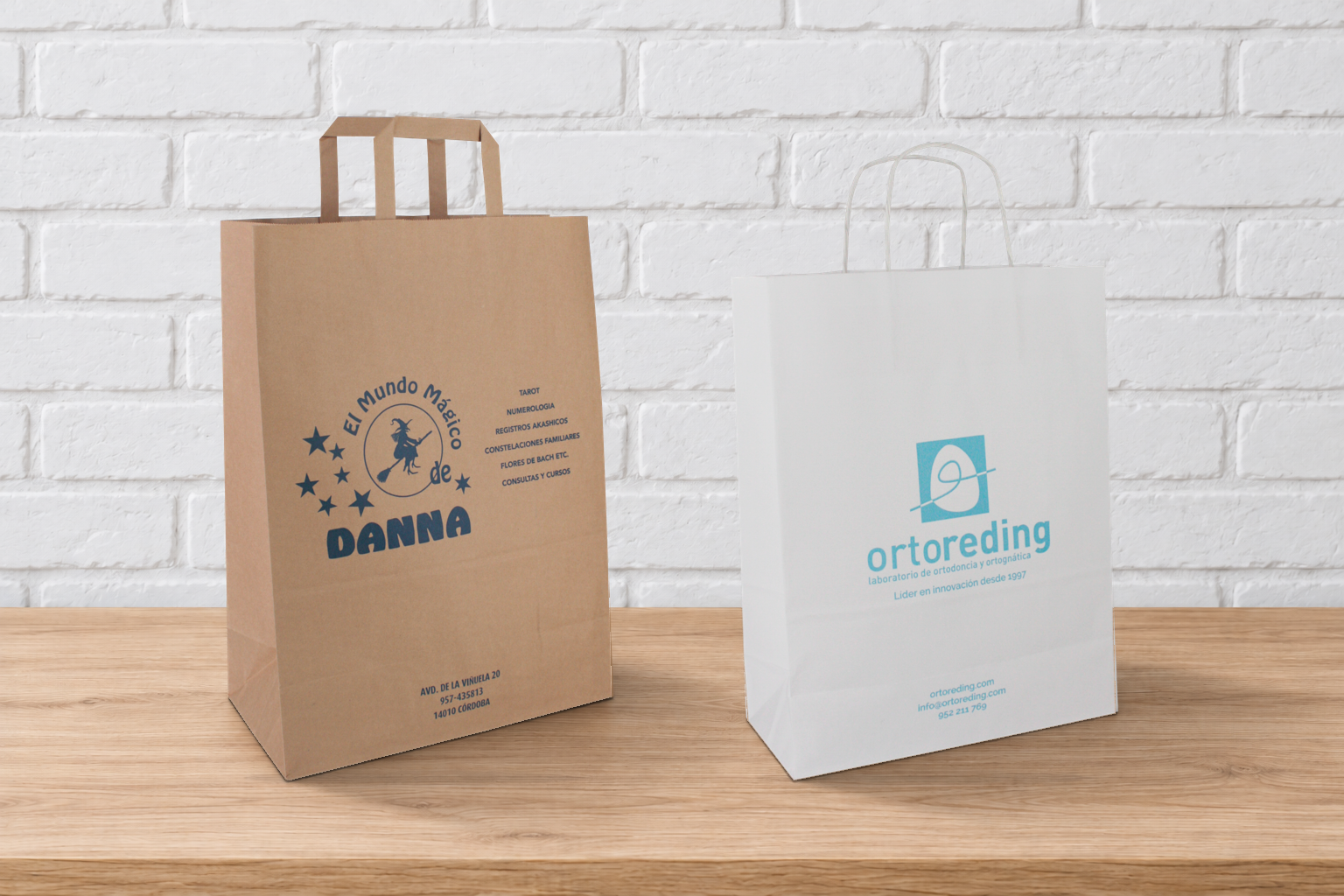Bolsas de papel kraft con asa plana y blanca con asa rizada, personalizadas con logotipos y apoyadas sobre una mesa ante una pared de ladrillo blanco